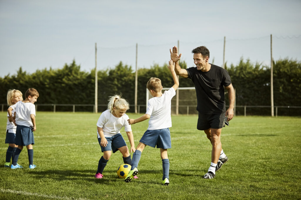 Smiling male soccer coach high-fiving with boy. Children are practicing on field. They are in sports uniforms.