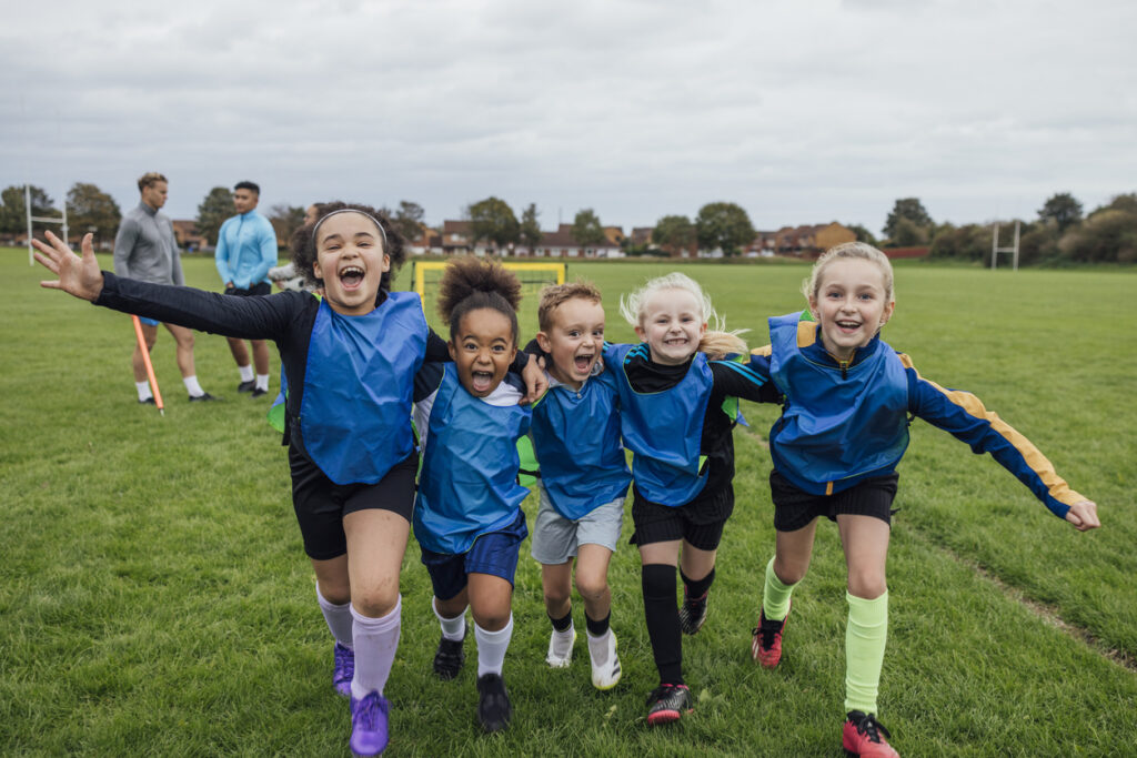 Front view of a small group of boys and girls wearing sports clothing, football boots and a sports bib on a football pitch in the North East of England. They are at football training where they are doing different football training drills. They are running and celebrating with their arms around each other in front of a football goal while smiling and looking at the camera.