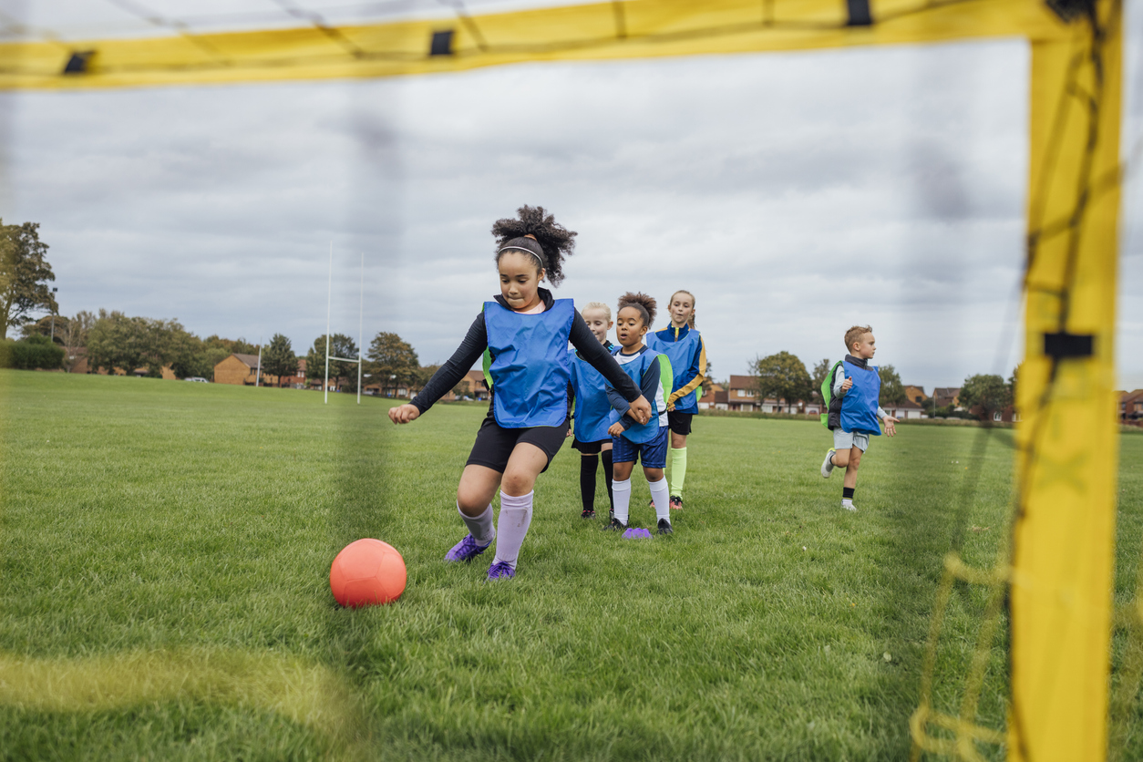 A wide shot of a group of children wearing sports clothing, football boots and a sports bib on a football pitch in the North East of England. They take turns shooting at the goal at football practice.